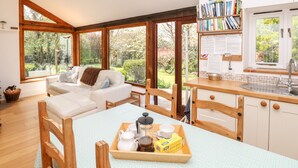 A cozy room with a sofa, a dining table with a tray of breakfast items, and a kitchen area with a sink and bookshelves. - Puffin Cottage (Axminster)