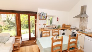 A compact kitchen with a dining area, a built-in oven, a washing machine, and a large window overlooking a garden. - Puffin Cottage (Axminster)