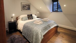 A bedroom with a sloped ceiling, a bed with a blue quilt, two bedside lamps, a framed picture on the wall, and a window with a view of trees. - Lake View (Windermere)