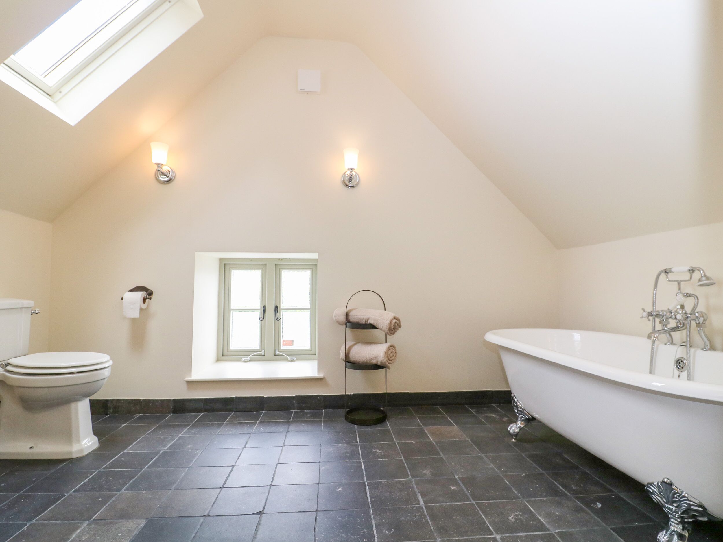 Ein Badezimmer mit schräg versiegelter Decke, einer Badewanne, einer Toilette, einem Fenster und zwei Wandleuchten.