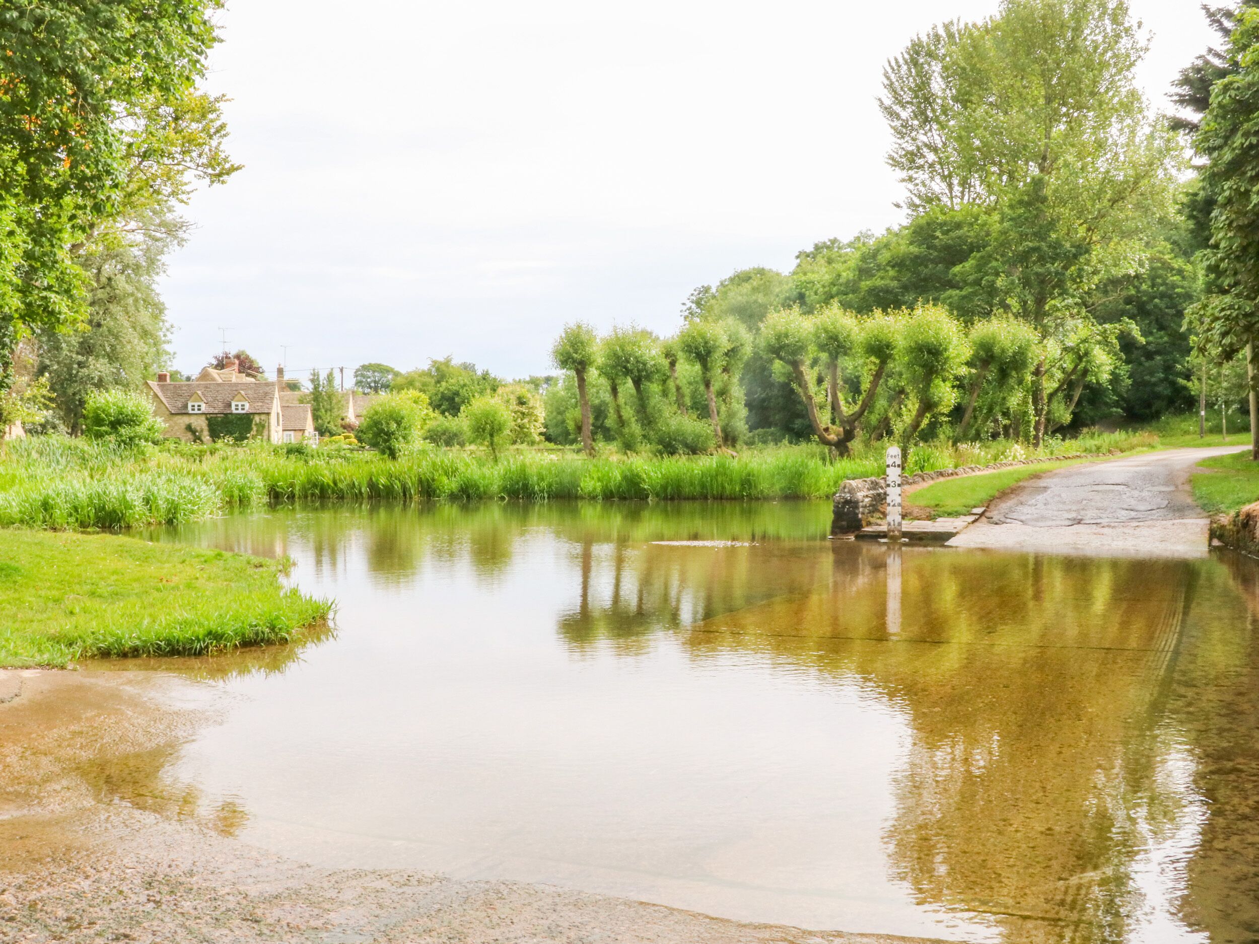 A calm river with a bridge, surrounded by lush greenery and a few buildings in the background.