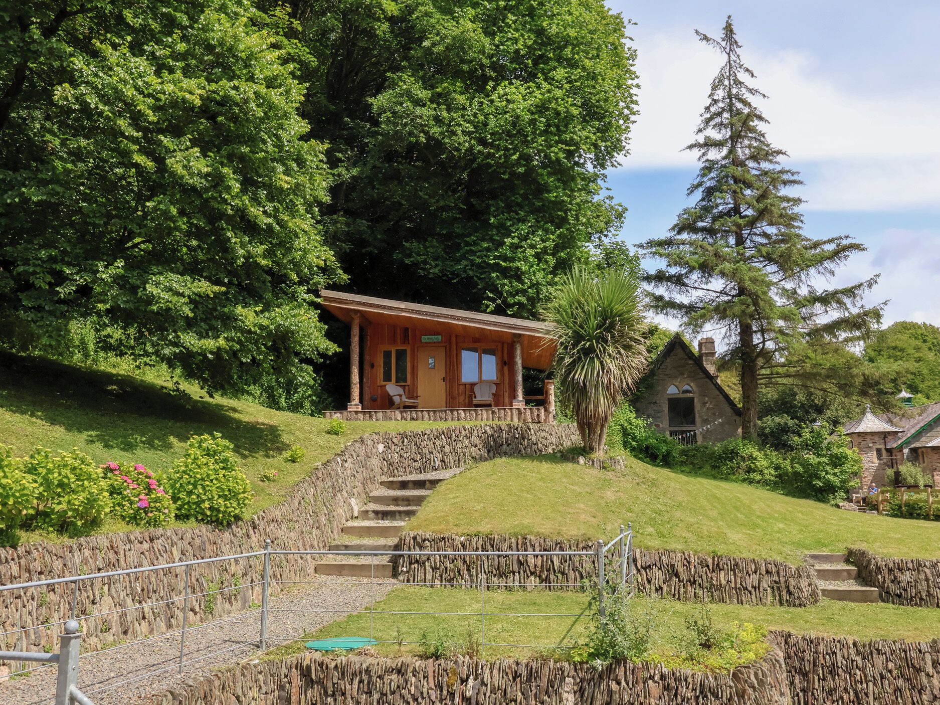 Ein rustikales Chalet mit einer Steintreppe, die zu einer Holzveranda führt, umgeben von üppiger Vegetation und einer hohen Nadelbaum.