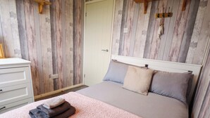 Une chambre avec un mur en bois, un lit avec une literie grise, une commode blanche et un tapis rose.
