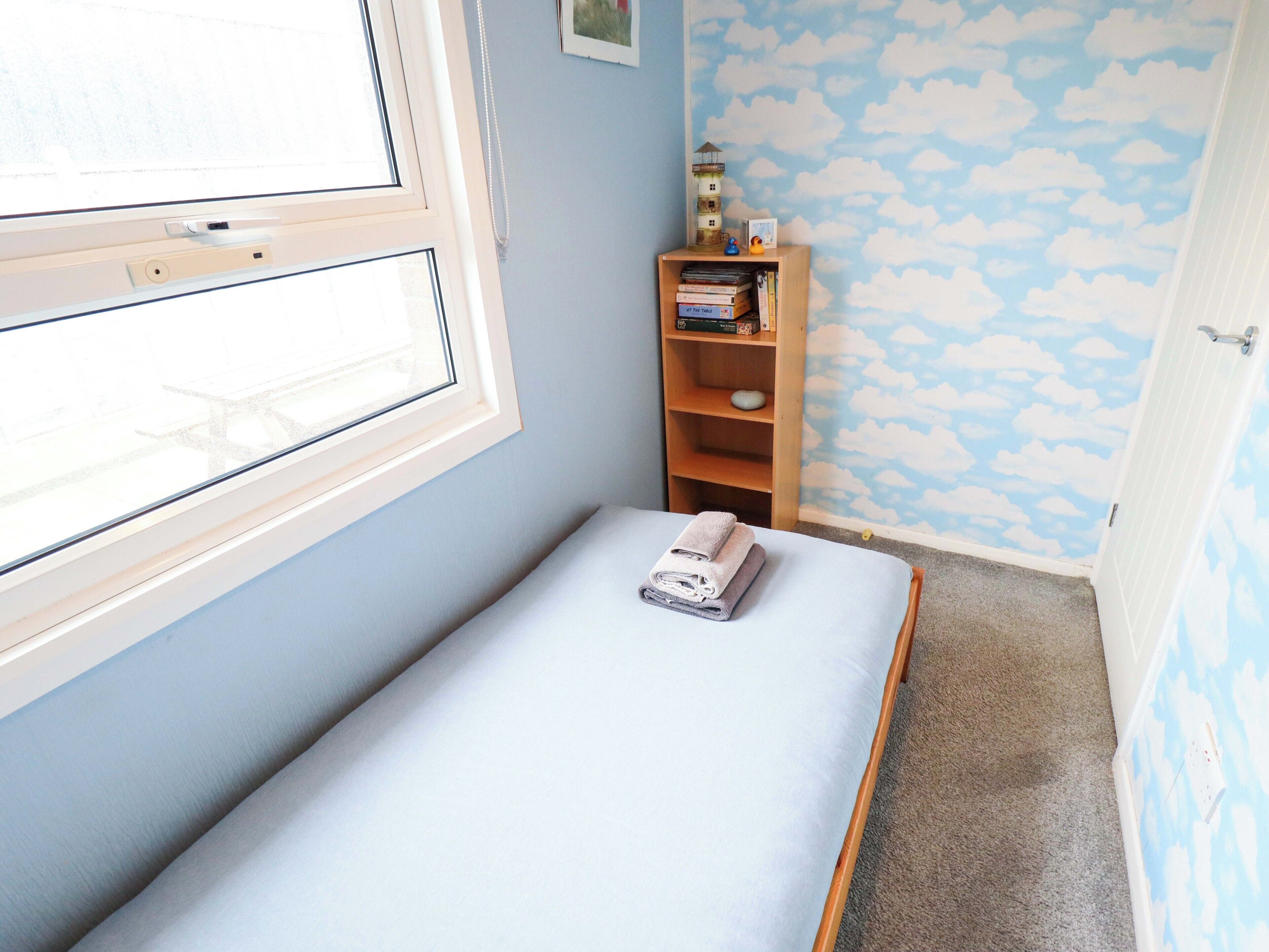 Une petite pièce avec un lit simple, une fenêtre, une bibliothèque et un mur orné d’un motif de nuages.