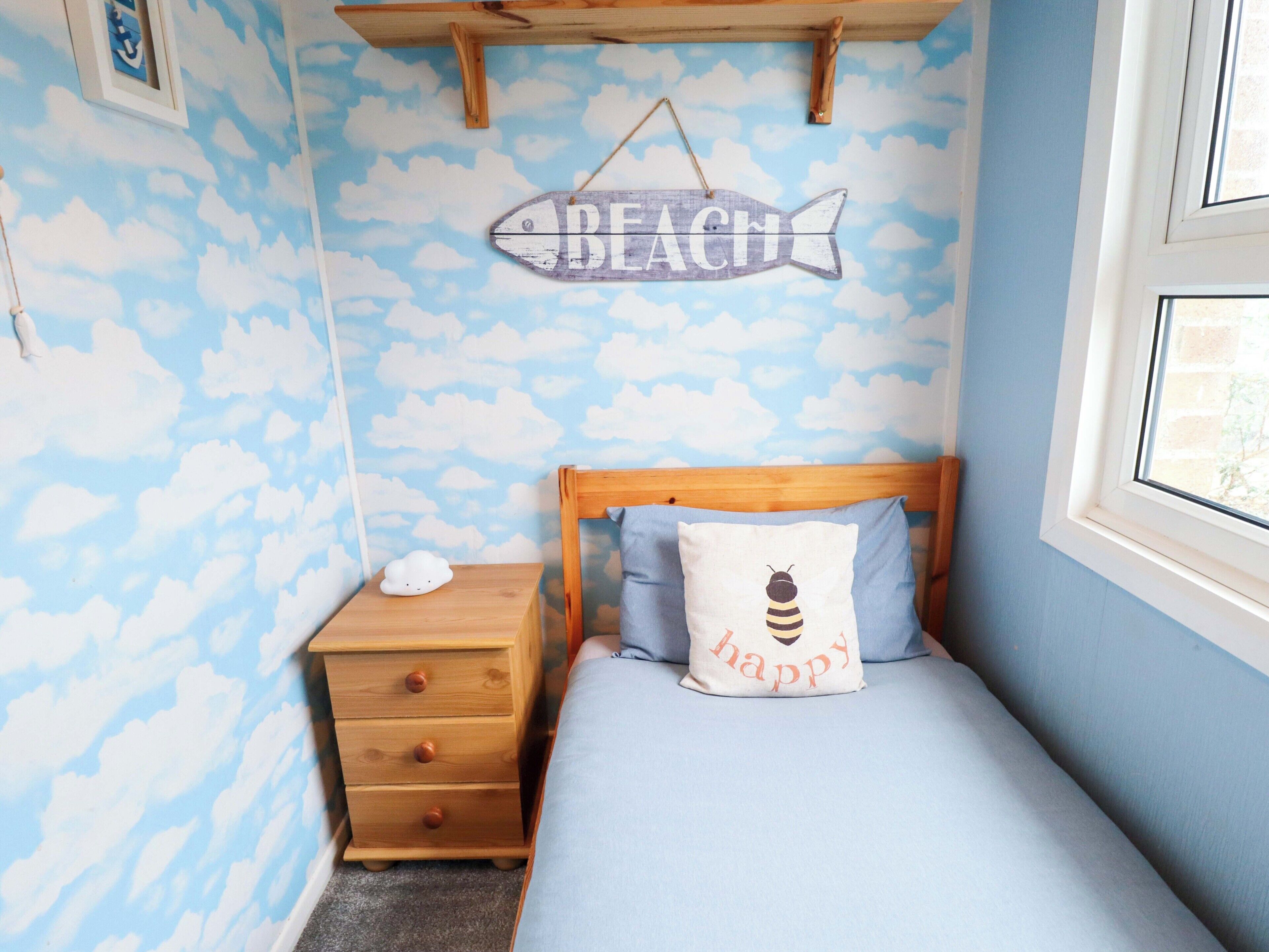 Une chambre avec un lit, une table de chevet, une commode en bois, un mur orné d’un motif de nuages et un panneau indiquant « PLAGE ».