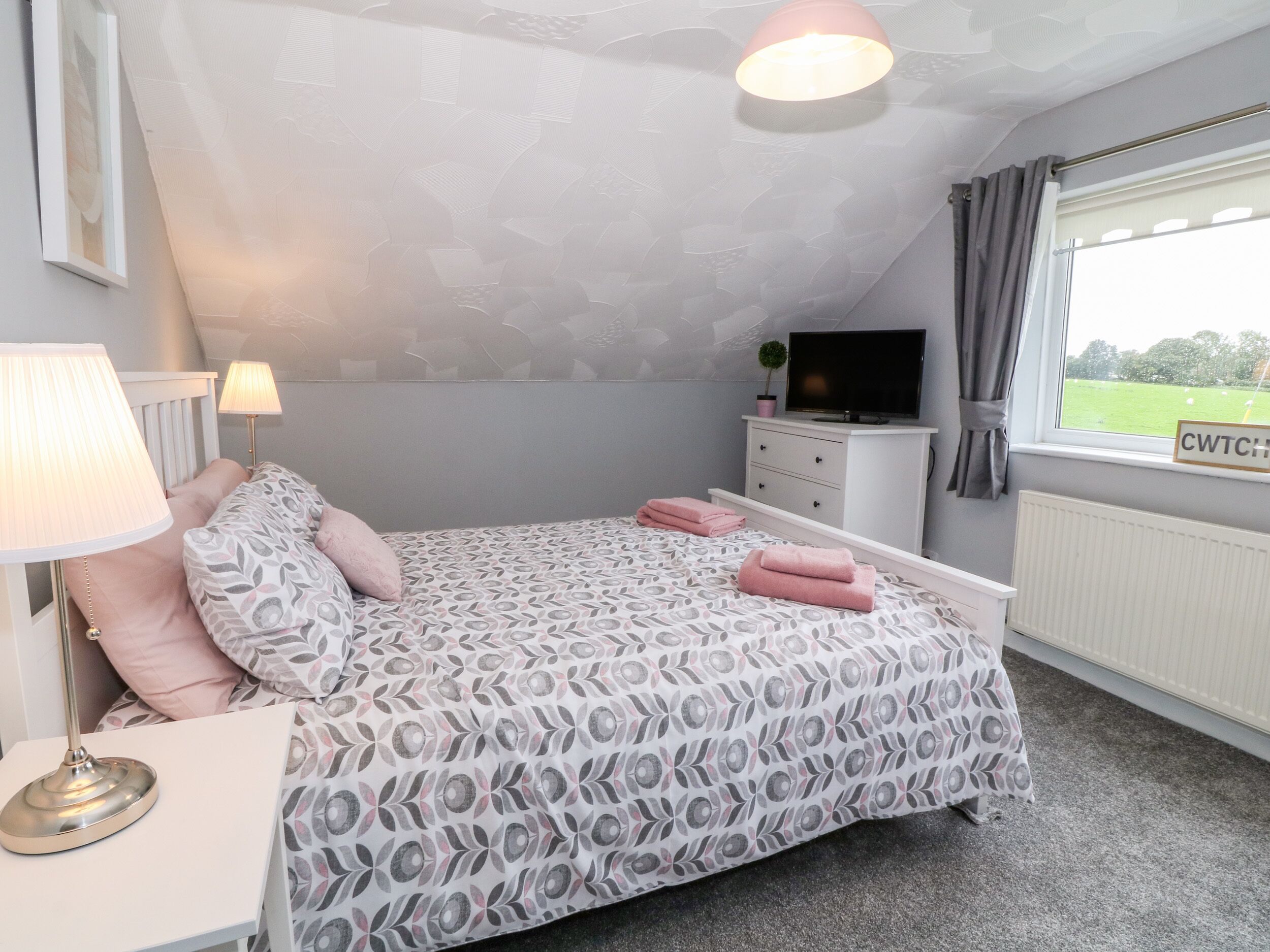 A bedroom with a bed, bedside lamps, a dresser, a television, and a window with a view of a green field.
