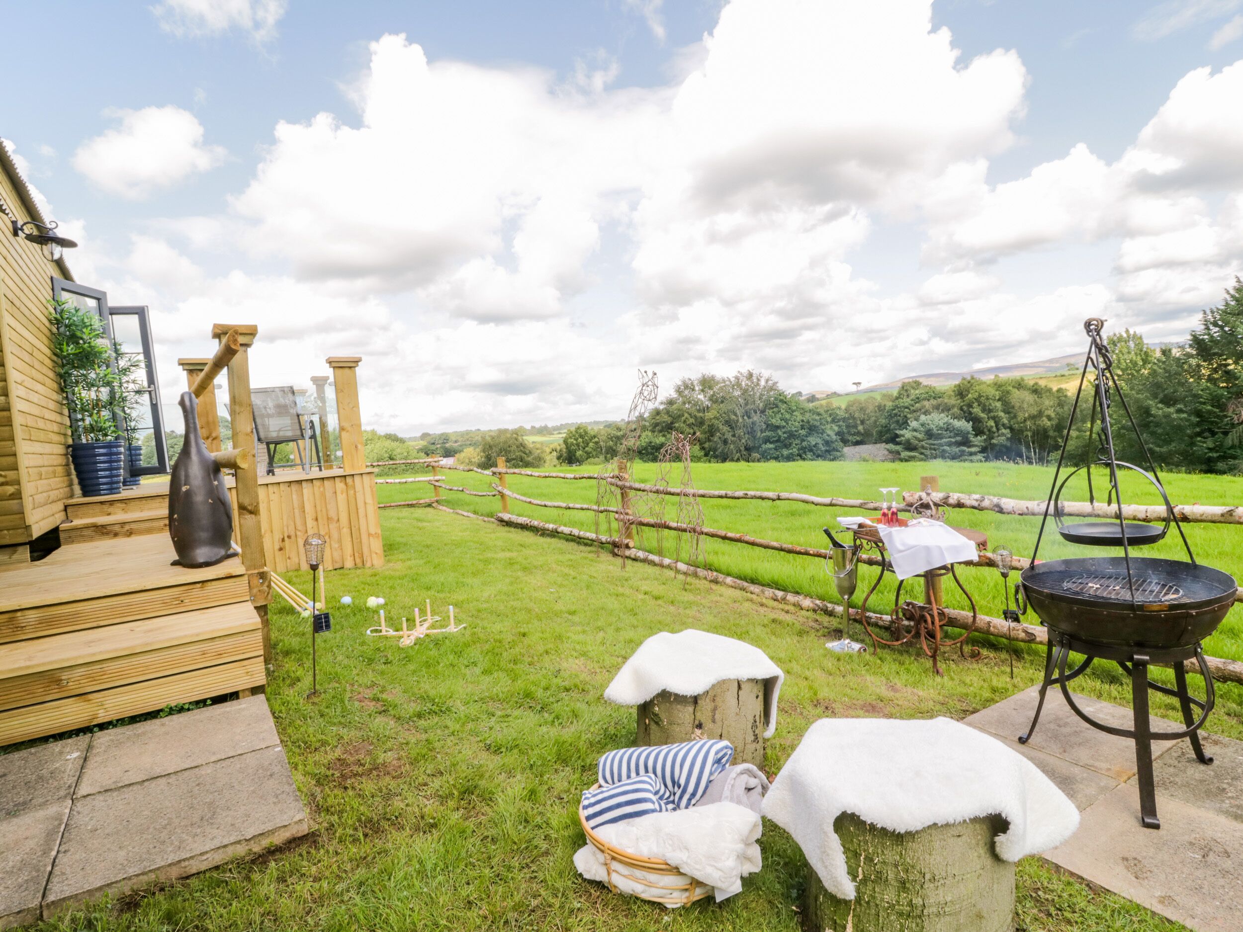 Een rustieke buitenomgeving met een houten terras, een grote zwarte barbecue en een omheind gebied met een grasveld op de achtergrond.