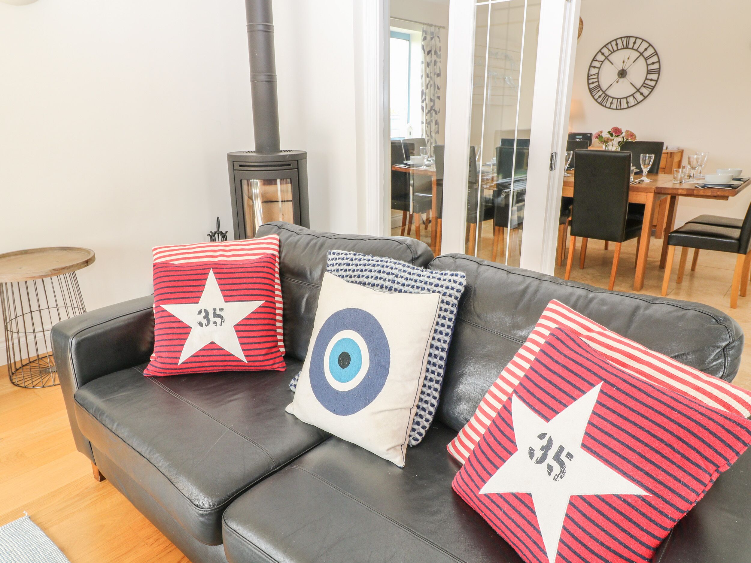 Een woonkamer met een zwarte leren bank, drie kussens met sterrenmotieven, een houten tafel en een klok aan de muur.