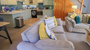 A spacious living area with a sofa, a dining table, and a kitchen with a washing machine. - Waterhead Cottage (Windermere)