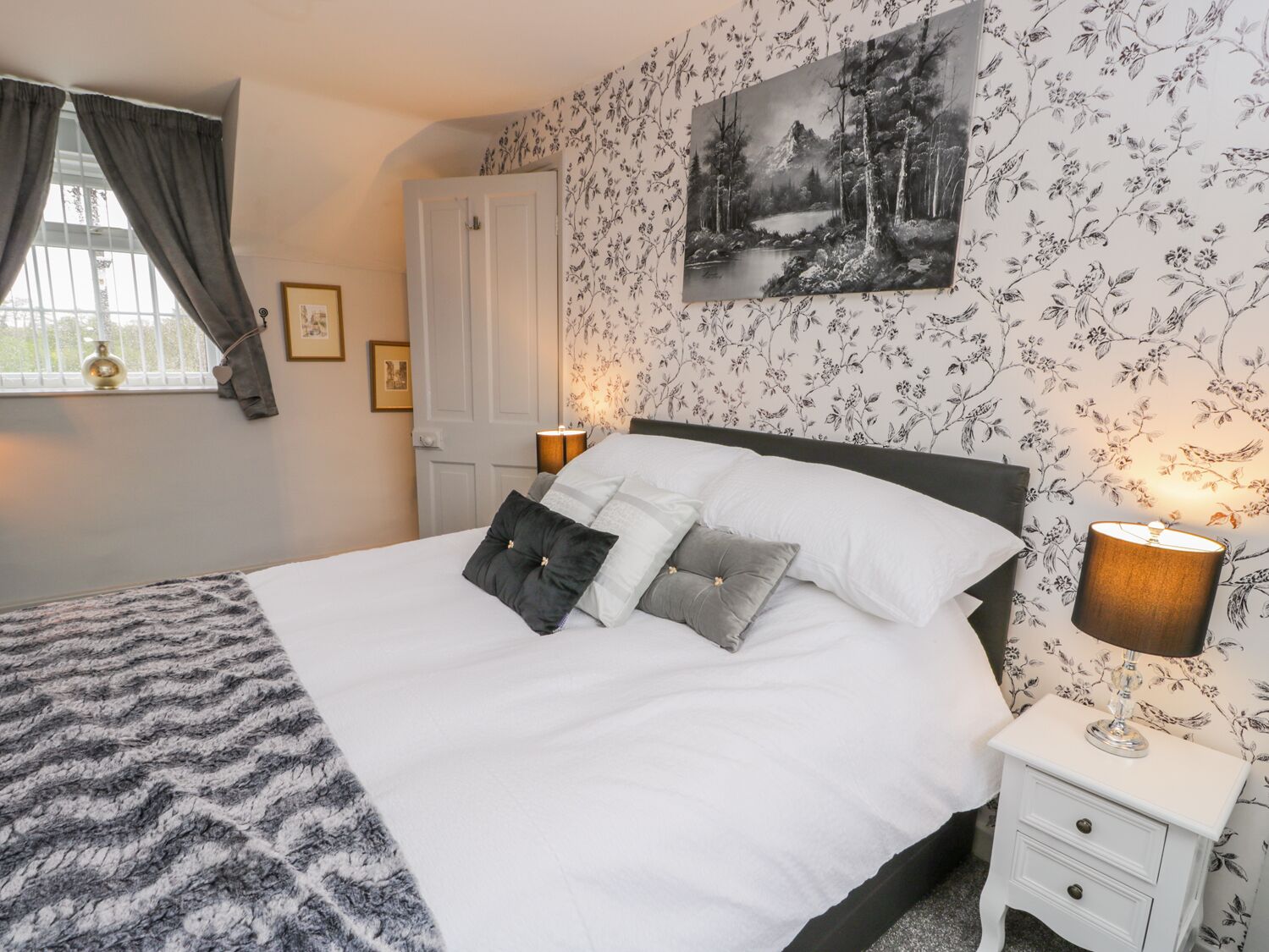 Een slaapkamer met een bed, twee nachtkastjes met lampen, een raam met gordijnen, een deur en bloemenbehang.