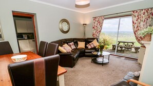 A living room with a brown leather sofa, a wooden dining table, and a view of the outdoors through a glass door.