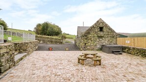 A stone building with a triangular roof, a stone wall, and a paved patio with a wooden table and benches. - Tai Ucha (Corwen)