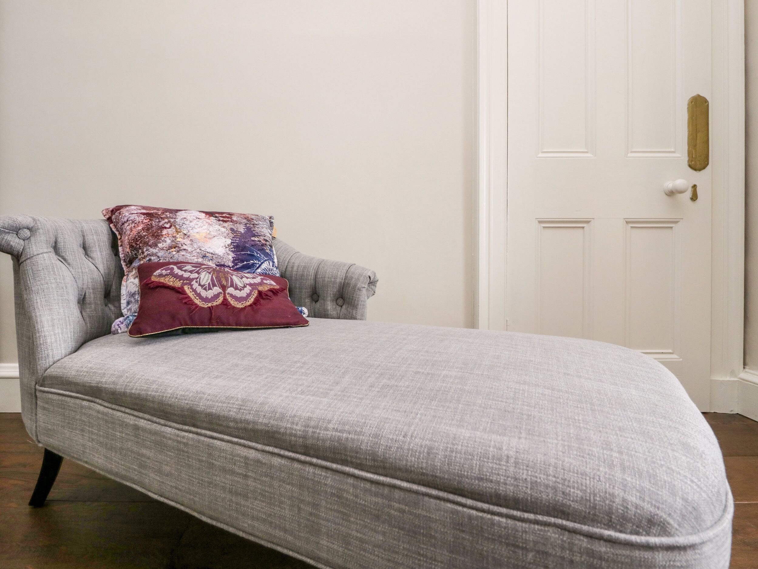 A grey upholstered sofa with a patterned cushion against a white wall and a white door.