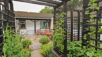 Un área de jardín cubierta con un pérgola de madera, un edificio blanco con techo de tejas y diversas plantas y flores.