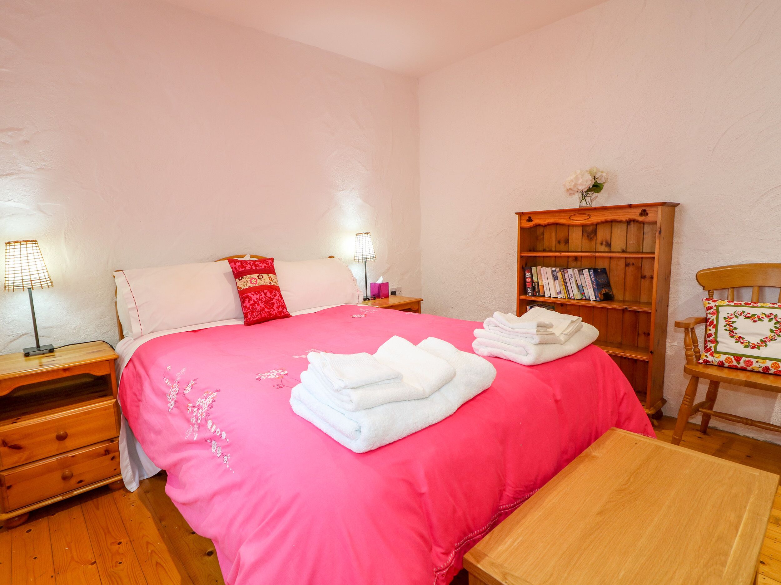 A bedroom with a large bed, a wooden nightstand with a lamp, a wooden bookshelf, and a wooden chair.