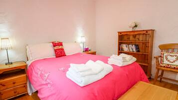 A bedroom with a large bed, a wooden nightstand with a lamp, a wooden bookshelf, and a wooden chair.