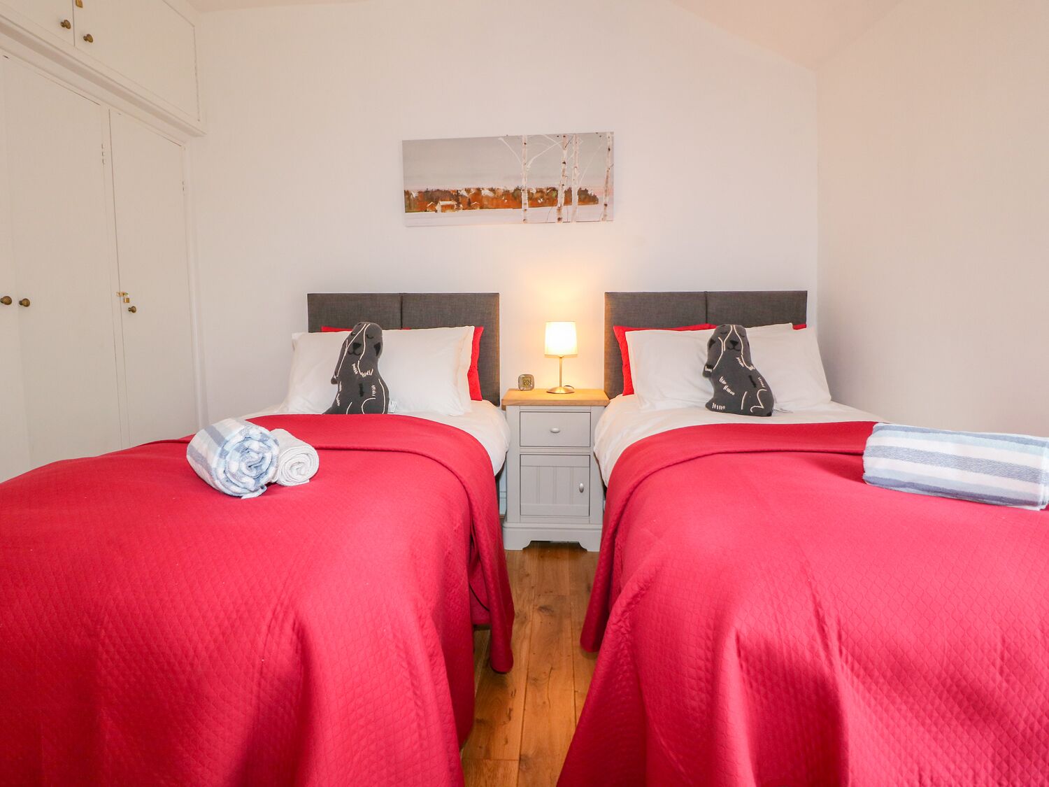 A hotel room with two beds, each with red bedding and a pillow featuring a black and white dog design. There is a small bedside table with a lamp and a clock.
