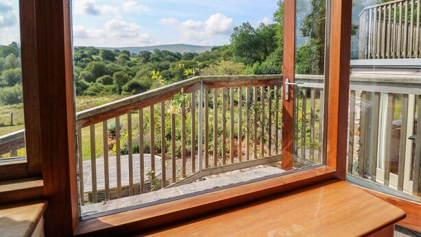 Un balcón de madera con puerta de cristal, con vistas a un jardín exuberante y colinas distantes.