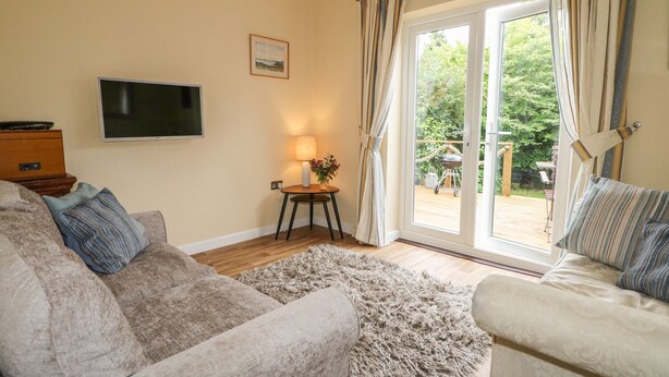 Ein Wohnzimmer mit einem beigen Sofa, einem gemusterten Sessel, einem Flachbildfernseher, einem kleinen Tisch mit einer Lampe und Blick auf einen Garten durch eine Glastür.