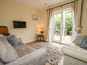A living room with a beige sofa, a patterned armchair, a flat-screen TV, a small table with a lamp, and a view of a garden through a glass door. - Beau Annexe (Ringwood)