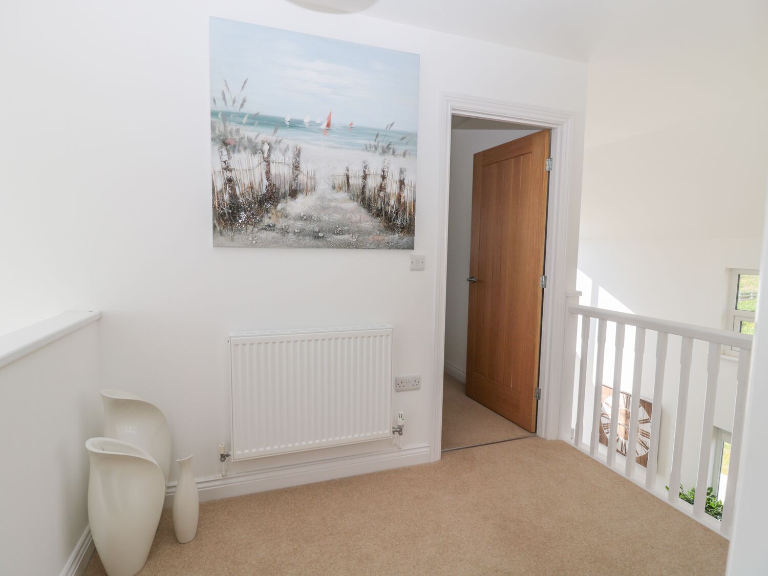 Een gang met een strandschilderij, een radiator en een houten deur die naar een andere kamer leidt.