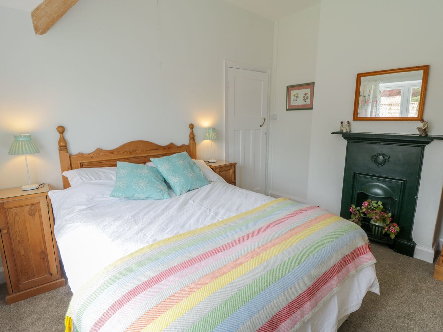A neatly made bed with a rainbow-striped blanket, two matching pillows, and a wooden headboard. There is a nightstand with a lamp and a dresser to the left. A green fireplace with a mirror above it is on the right side of the room.
