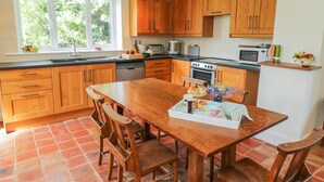 A kitchen with wooden cabinets, a dining table with chairs, a window with a view of greenery, and a countertop with various appliances. - Manor Cottage (Pickering)