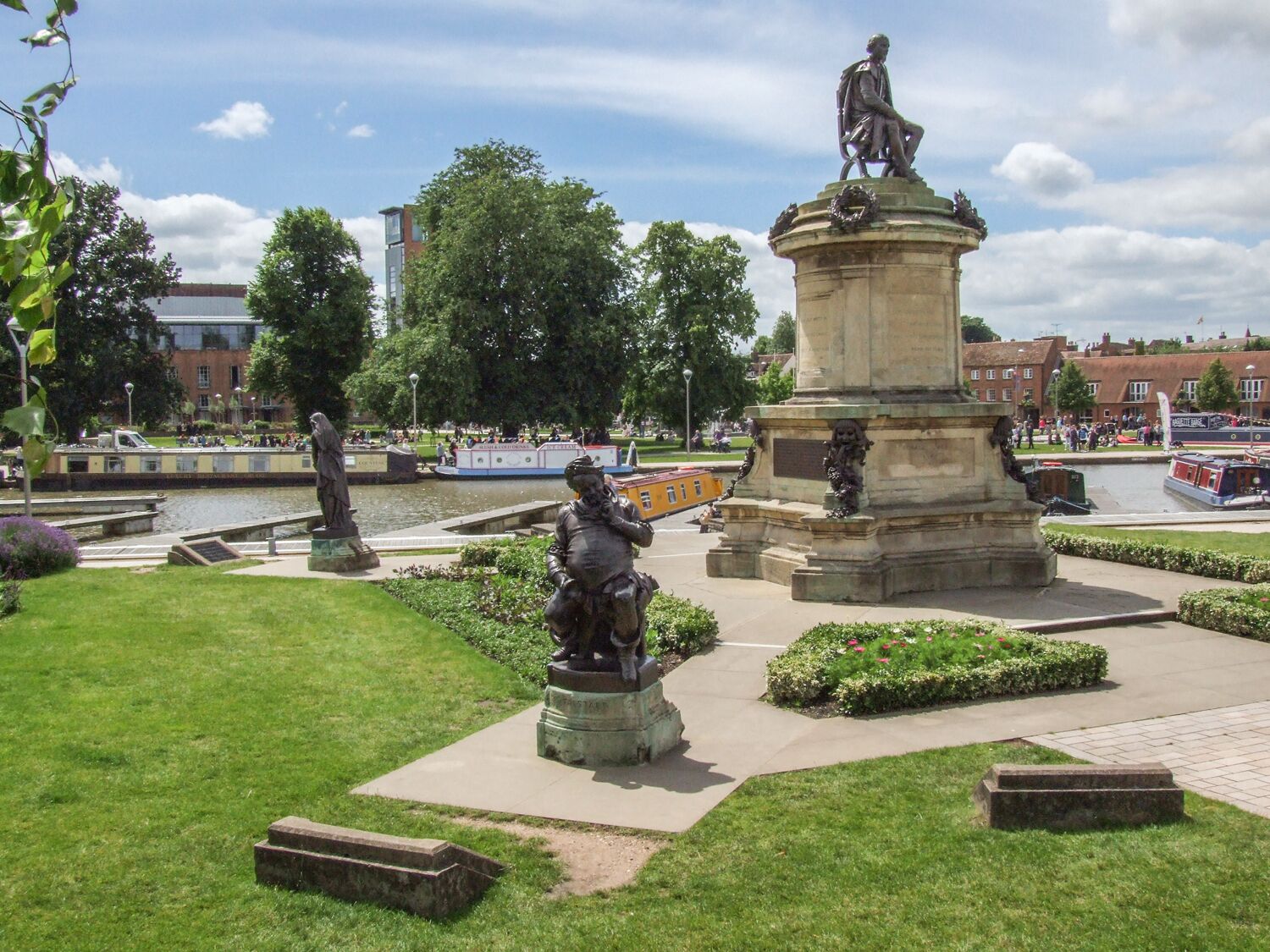 A public square with statues, a fountain, and a canal with boats.