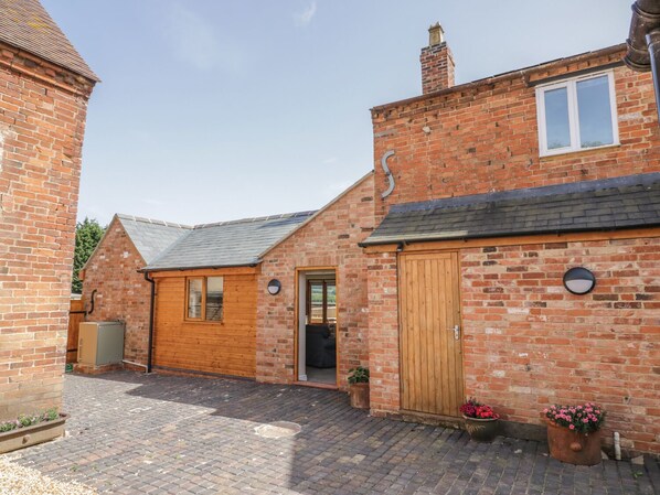 A brick house with a wooden door, a window, and a chimney. There is a paved courtyard with potted plants. - Cider Cottage (Stratford-upon-Avon)