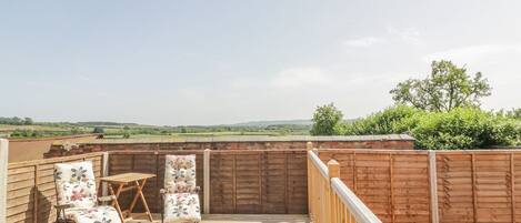 A wooden deck with two chairs and a table, surrounded by a wooden fence, overlooking a green landscape.