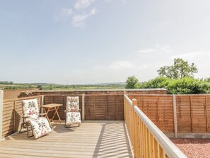 A wooden deck with two chairs and a table, surrounded by a wooden fence, overlooking a green landscape. - Cider Cottage (Stratford-upon-Avon)