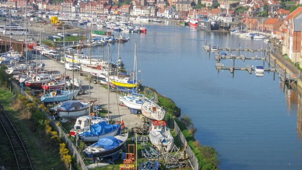 A marina with boats docked along the waterfront, surrounded by residential buildings and a historic building in the background. - Moor Croft (Whitby)