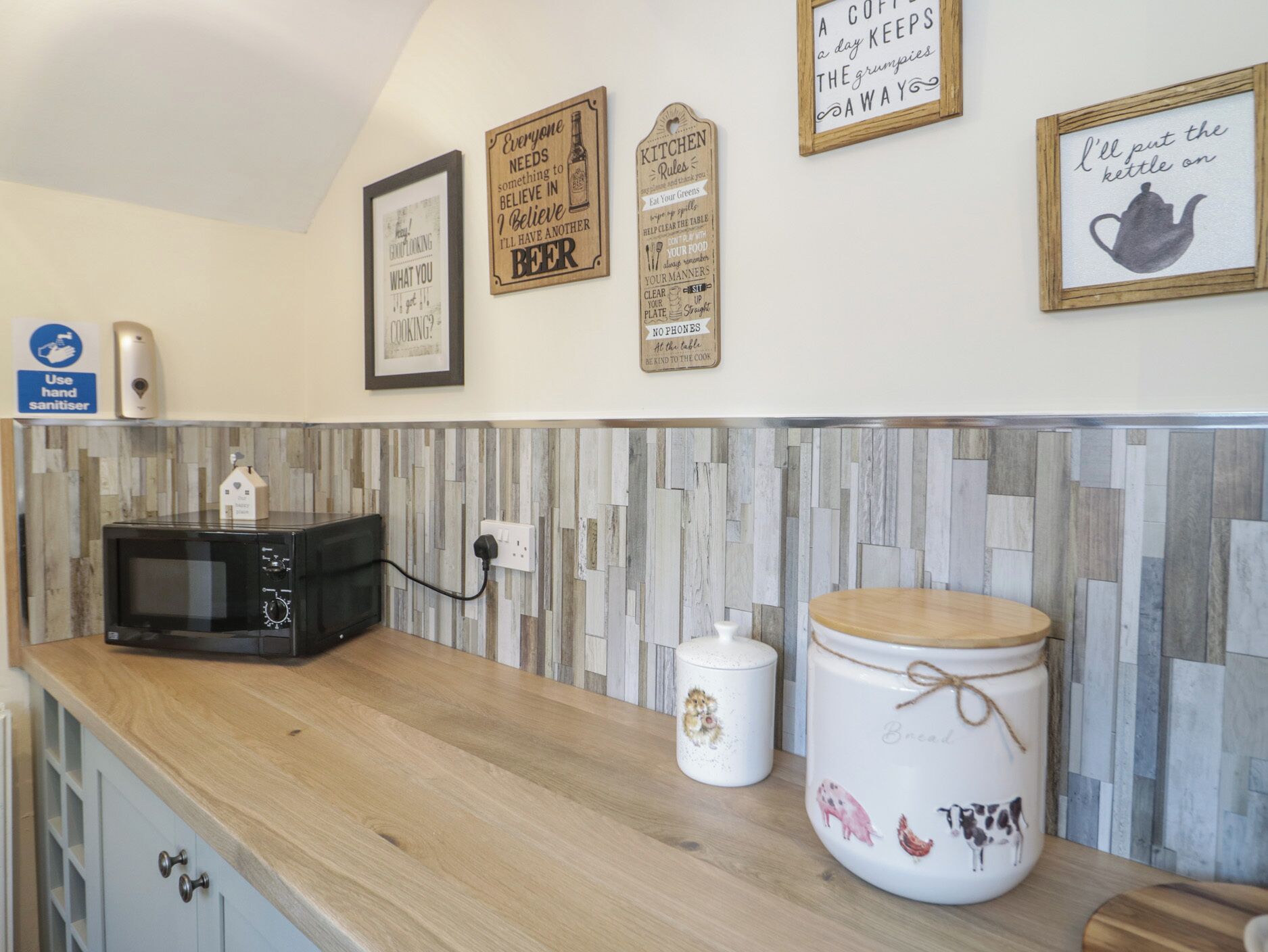 A kitchen with a microwave, a kettle, and a jar with a lid. There are wall decorations and a hand sanitizer dispenser.
