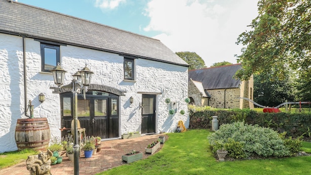 A white building with a stone facade, a large wooden barrel, and a well-maintained garden with a lawn and potted plants. - Primose Cottage (Milford Haven)