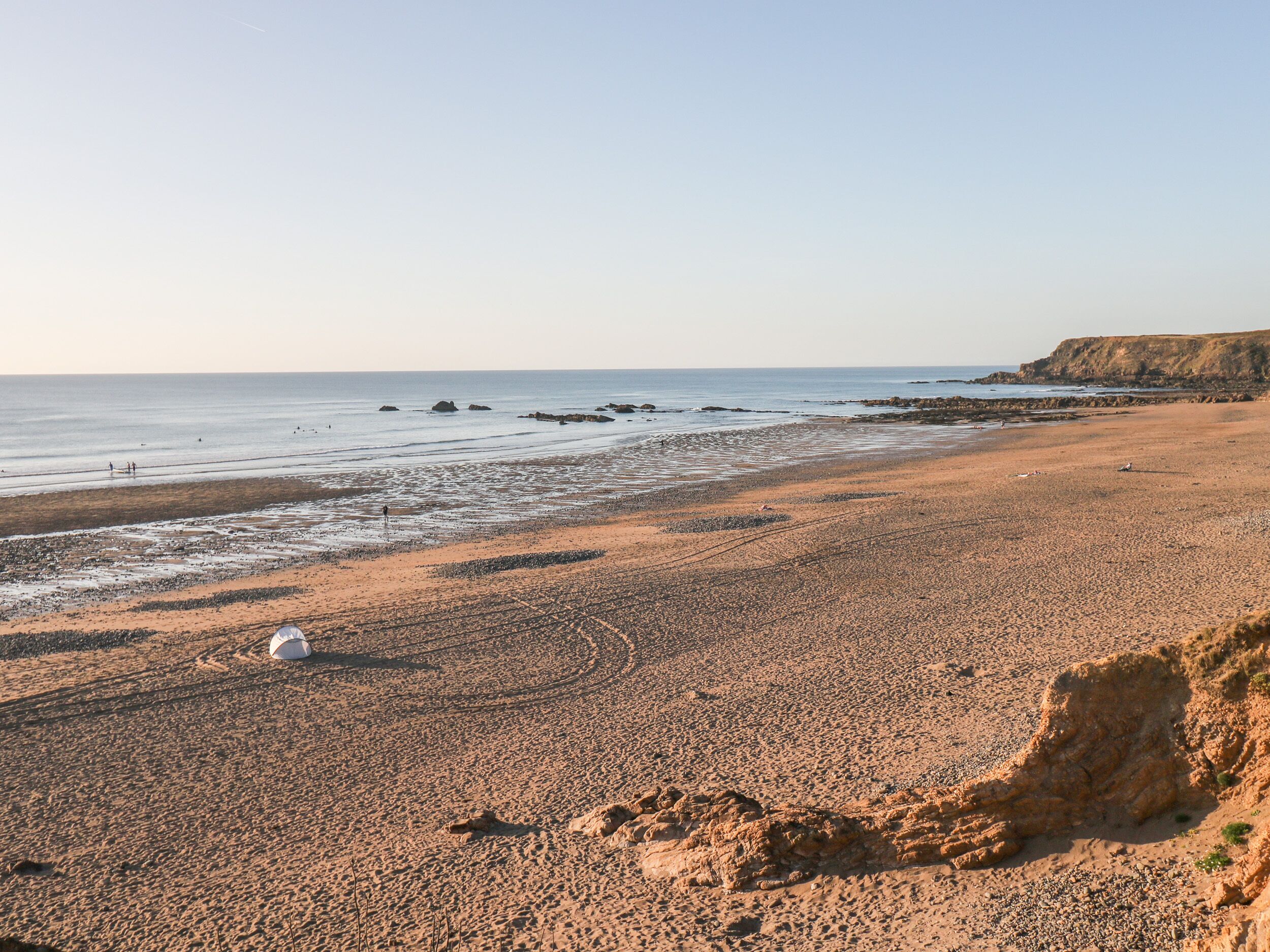 Una playa costera con cielo despejado, costa rocosa y acantilados al fondo.