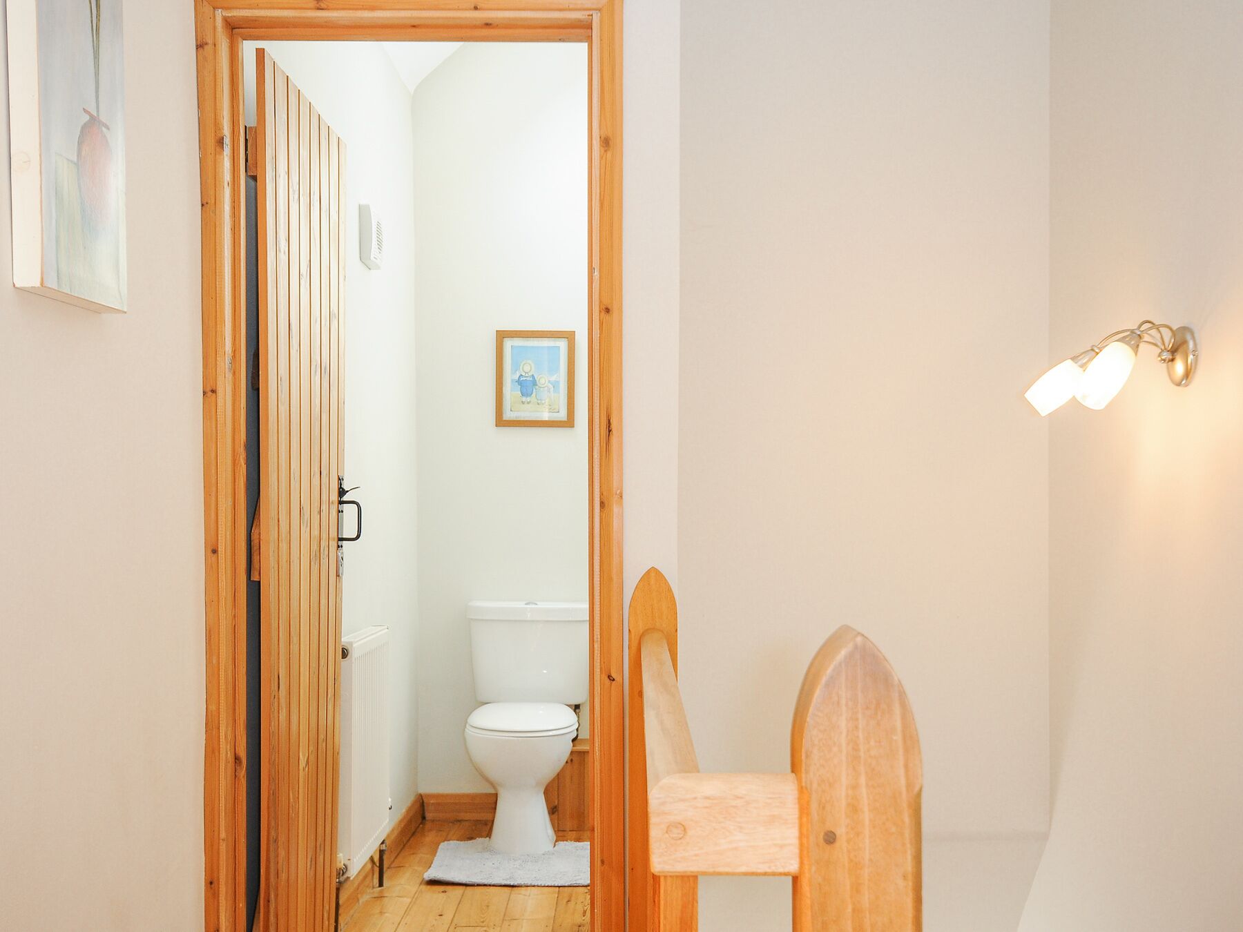 A narrow hallway leading to a bathroom with a white toilet, a wooden chair, and a framed picture on the wall.
