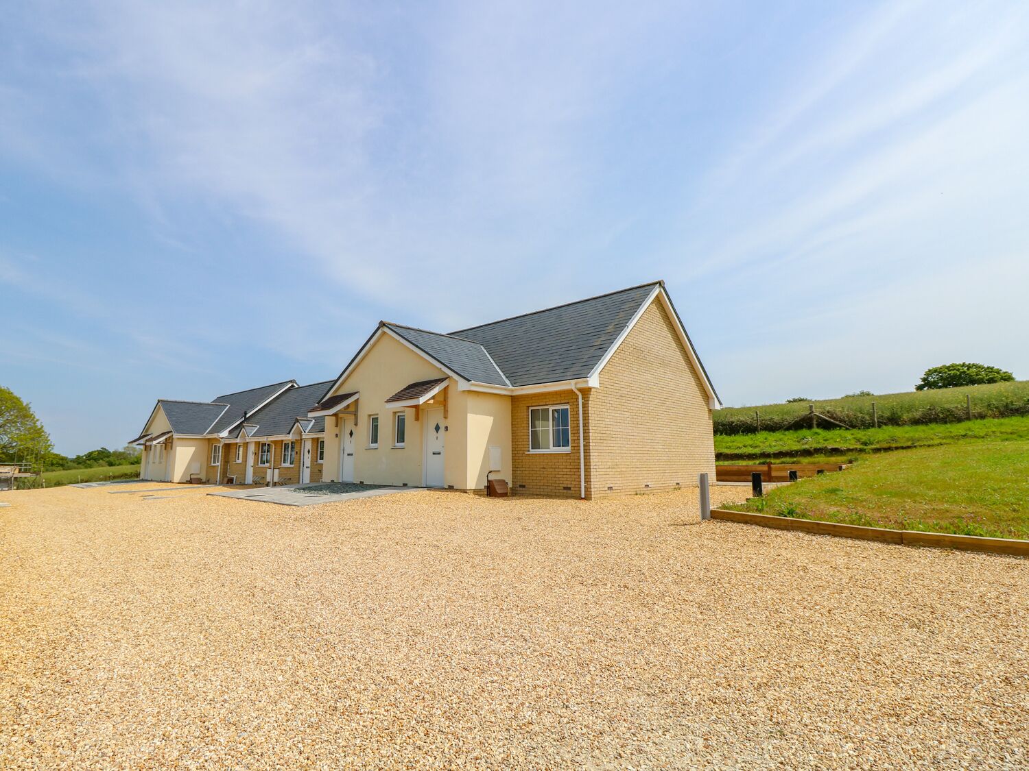 A row of single-story houses with a gravel driveway, surrounded by a grassy field and a fence.