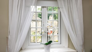 A window with white curtains, a potted plant, and a view of buildings outside. - Pemba Cottage (Skipton)