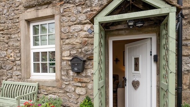 A stone building with a wooden bench, a potted plant with red flowers, and a door with a heart-shaped decoration. - Pemba Cottage (Skipton)
