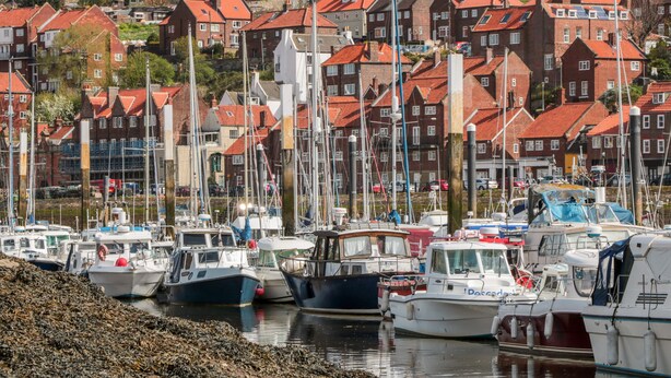 Un port où des bateaux sont amarrés, des bâtiments aux toits rouges et, au fond, un monument historique.