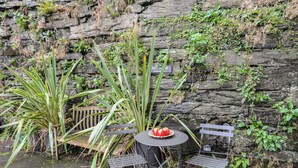 A stone wall with greenery, a metal table with chairs, and a basket of tomatoes. - The Old Cornhill (Porthmadog)