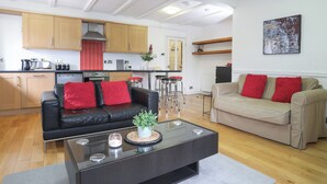 A modern living room with a black leather sofa and a beige sofa, both with red cushions. There is a glass coffee table with a plant and candles. The room has wooden flooring and a white ceiling with exposed beams. - The Old Cornhill (Porthmadog)