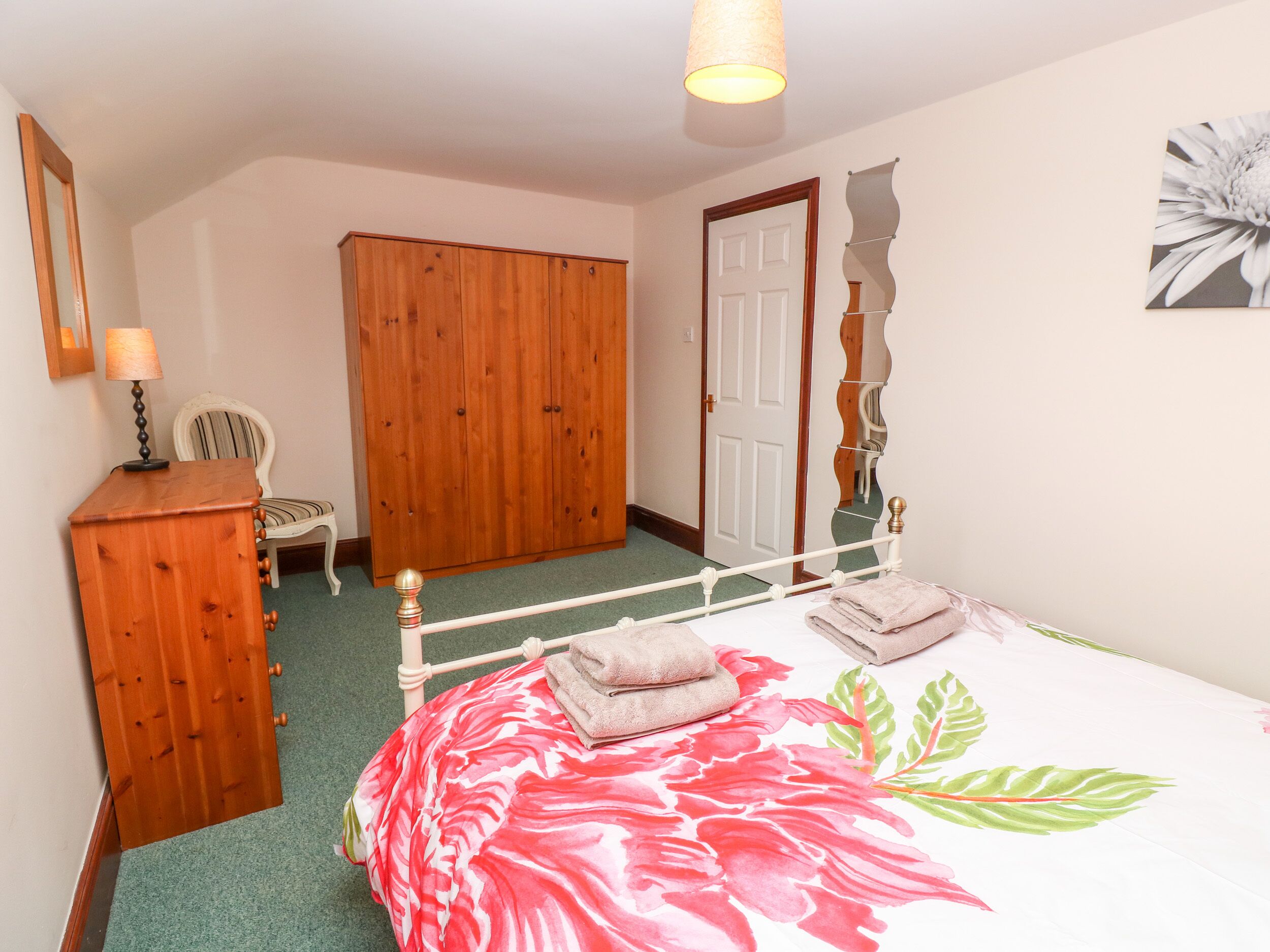 A bedroom with a bed, a wooden wardrobe, a chair, a door, and a wall-mounted mirror.
