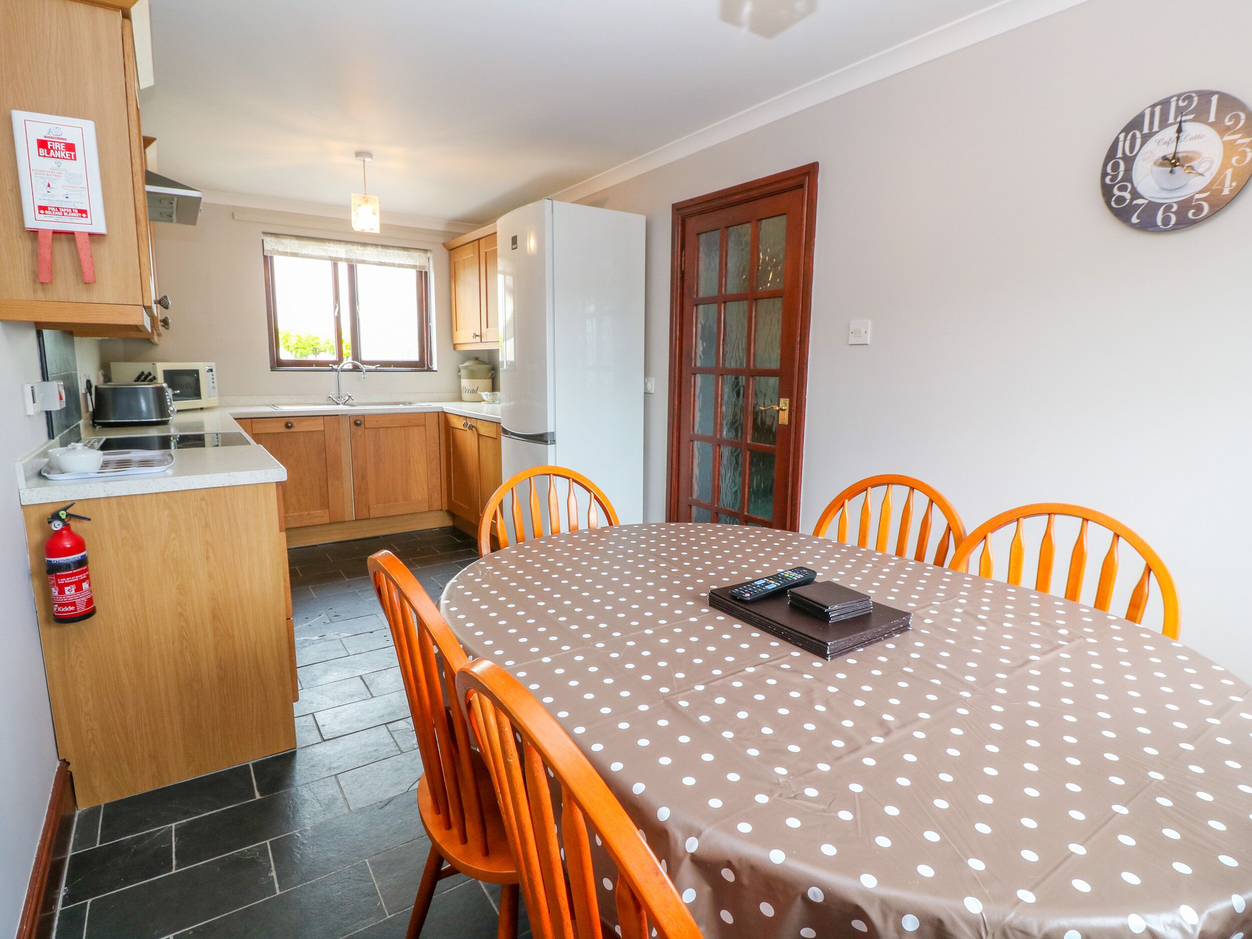A dining area with a polka dot tablecloth, wooden chairs, a clock on the wall, and a kitchen in the background.