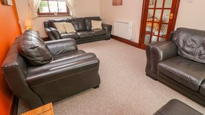 A living room with leather sofas, a radiator, a window with curtains, and a lamp. - Mossley Cottage (Penzance)