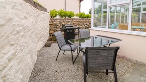 A patio area with a glass table and two chairs, a stone wall, and a view through a window. - Mossley Cottage (Penzance)