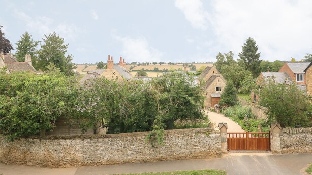 A stone wall with a wooden gate, leading to a rural area with houses and trees. - Wren Cottage (Oakham)