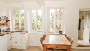 A kitchen with white cabinets, a wooden table, and a window overlooking a garden. - Wren Cottage (Oakham)