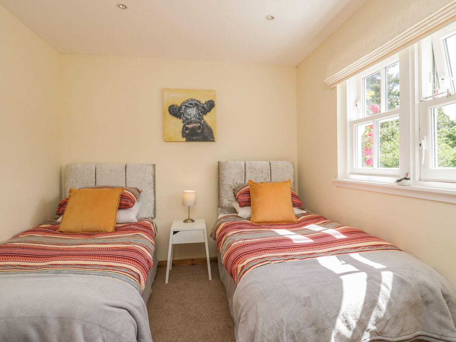 Een slaapkamer met twee bedden, een klein nachtkastje met een lamp, een schilderij aan de muur, en een raam met uitzicht op groen.