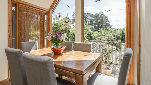 A dining area with a wooden table and grey chairs, a vase with flowers, and a view of greenery and birds outside. - Drumhead Bothy (Banchory)